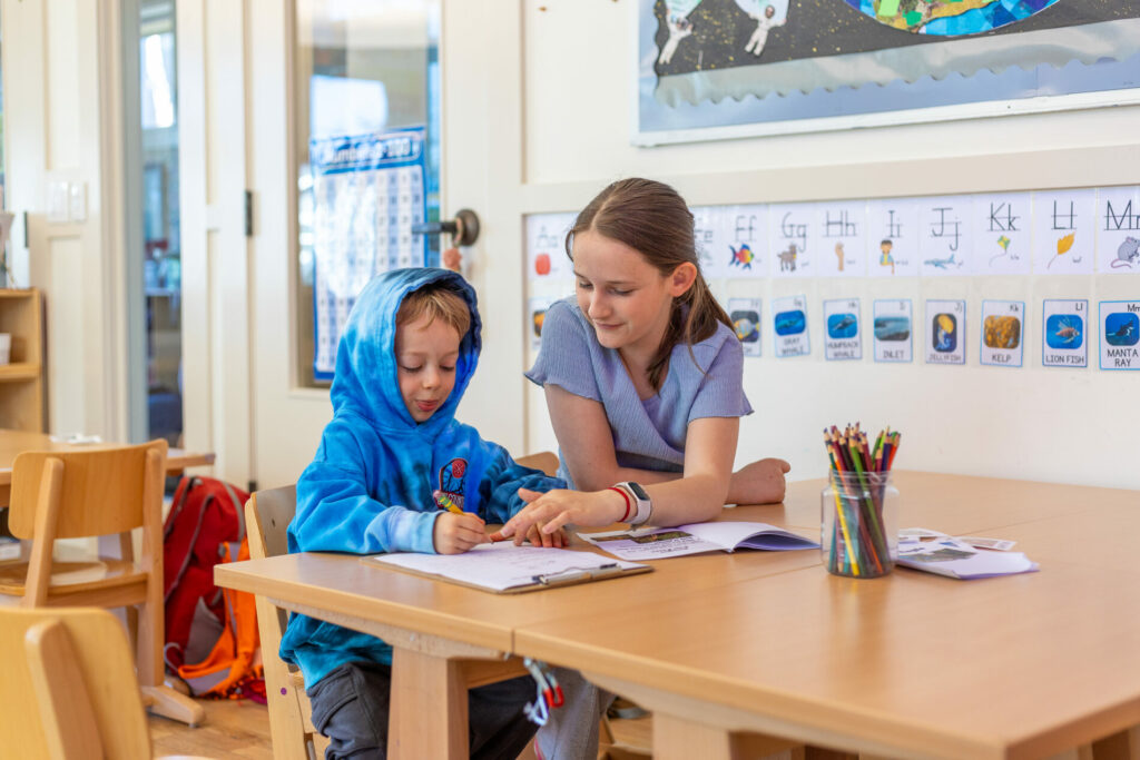 A fifth grade student helps a prekindergartner with his project at The Meridian School, serving grades pre-k–5th grade.
